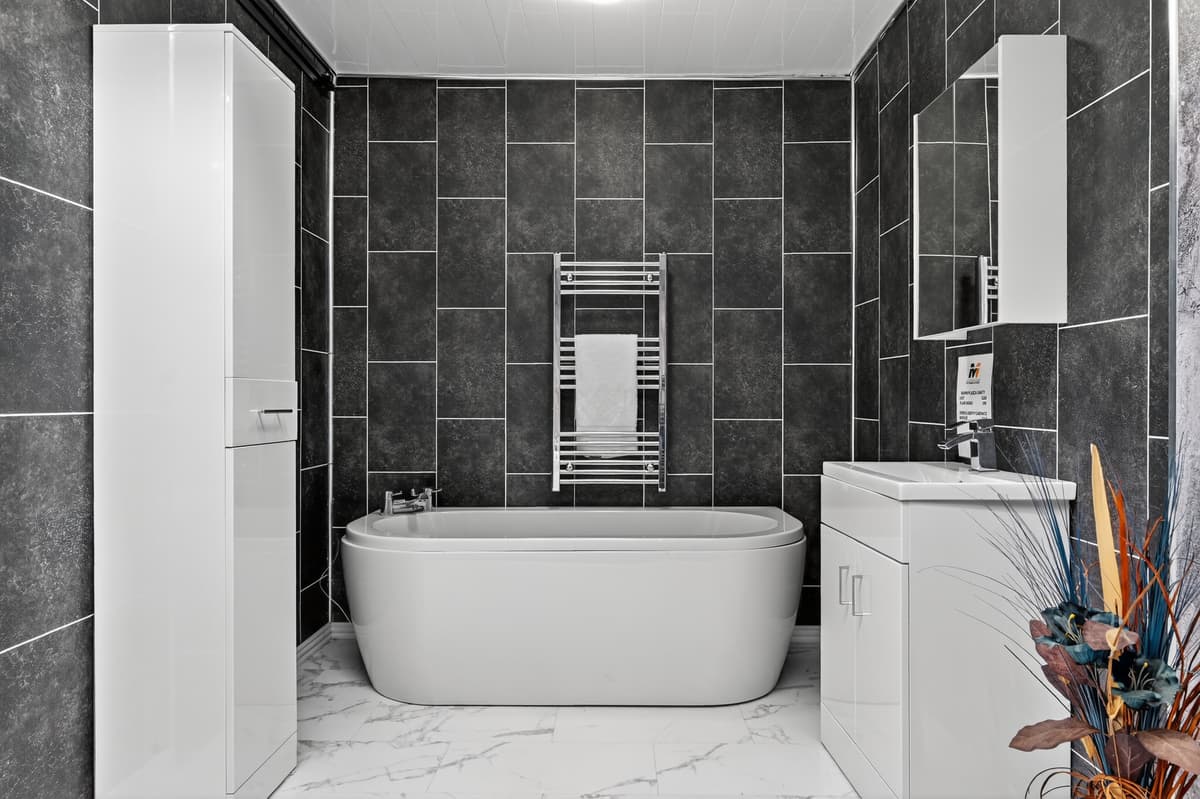 Modern bathroom with dark grey wall tiles, freestanding bath, white vanity unit and chrome heated towel rail at M1 Bathrooms Belfast.