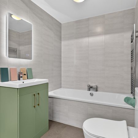 Sage green floor standing vanity unit with gold handles white bath light grey wall tiles chrome heated towel rail and LED mirror display in M1 Bathrooms Belfast showroom